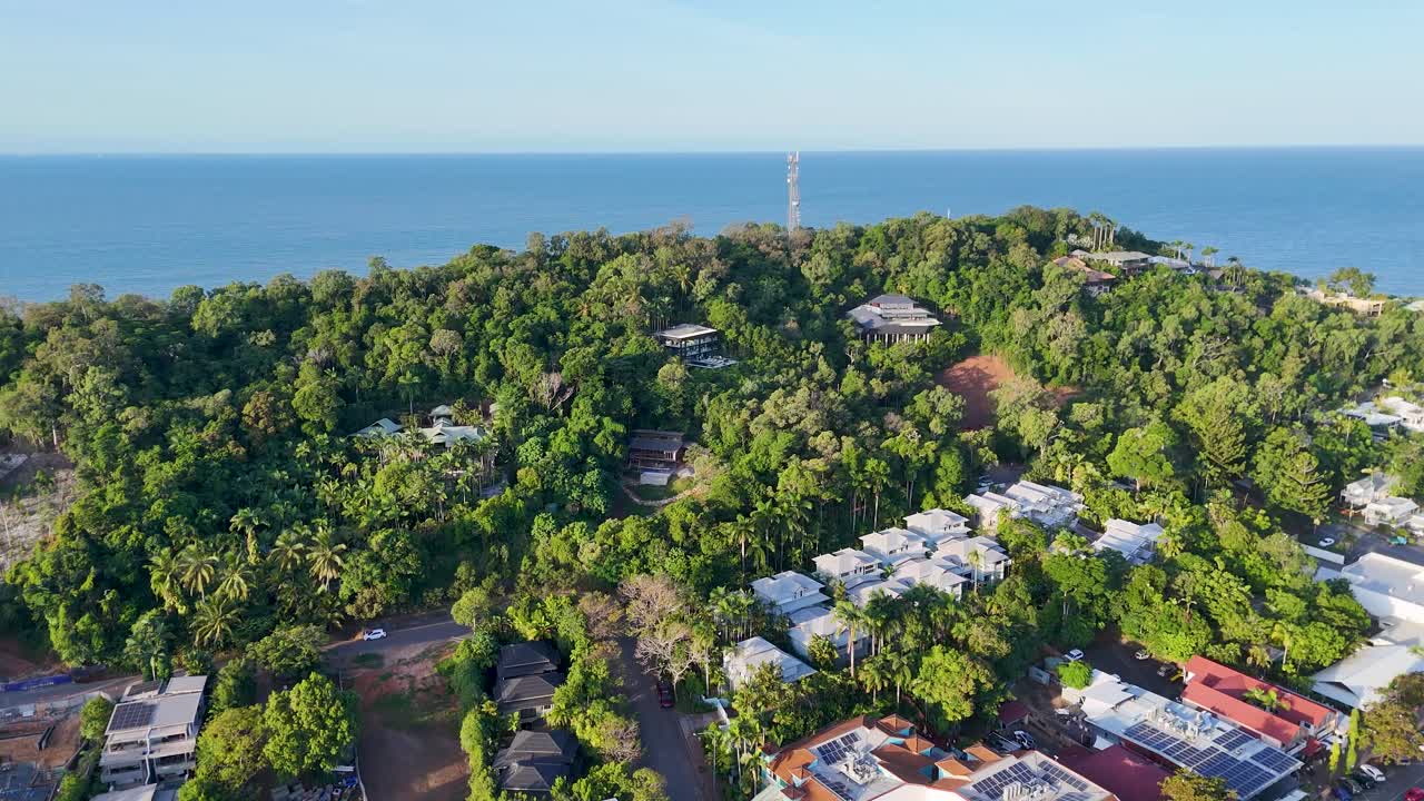 Drone footage captures a lush tropical town with vibrant greenery and coastal views under bright daylight