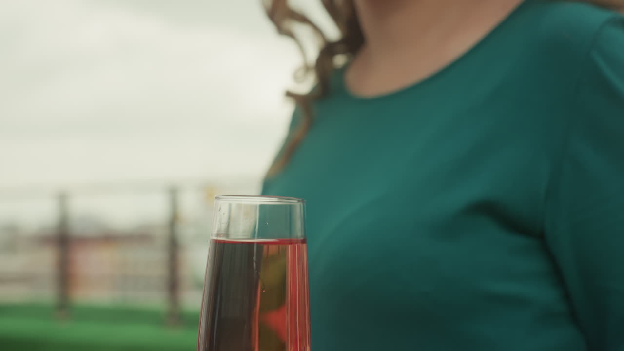 Brindisi su un tetto con spumante rosato con donna caucasica e uomo che offre un flute di vino rosato, mentre la donna in abito verde accetta il bicchiere, primo piano sulle mani e sullo stelo, sorriso delicato, ritratto spontaneo, skyline della città e sera