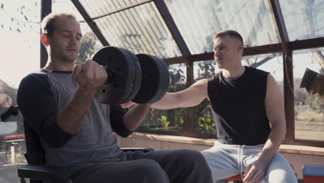 Hombre-Discapacitado-En-Silla-De-Ruedas-Haciendo-Ejercicio-Con-Pesas-En-Casa-Mientras-Su-Entrenador-Personal-Lo-Ayuda