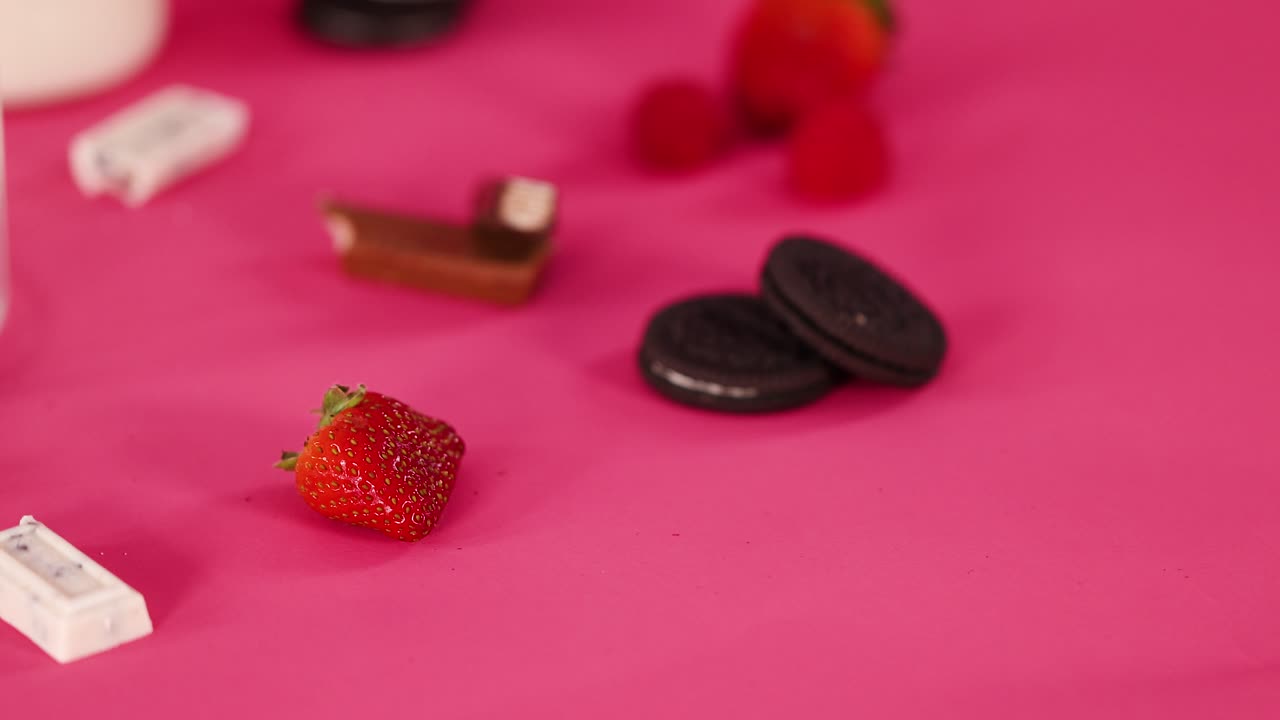 Strawberries, raspberries, cookies, and chocolate bars move on vibrant pink surface under soft lighting