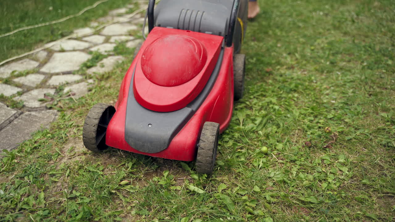 Red lawn mower. Electric mower machine trimming grass in the garden of private territory. Woman pushing lawn equipment.