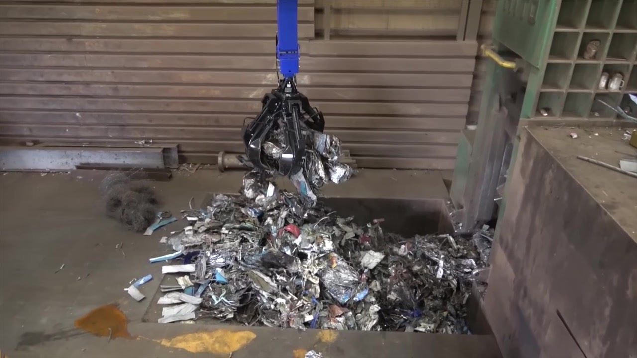 Blue orange peel grapple lifting crushed scrap metal from a pile inside an industrial recycling sorting warehouse