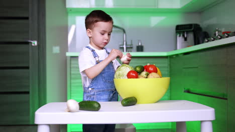 cute baby boy in jeans romper stands at the table in the kitchen. lovely toddler takes vegetables from the table and puts them into a big bowl.