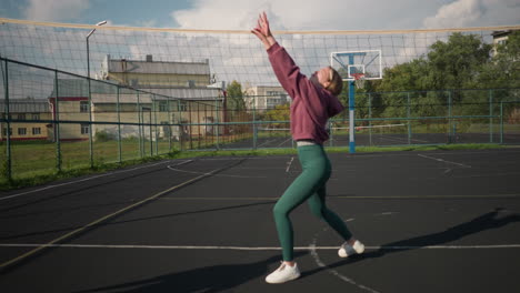 female athlete in volleyball training session passes ball to coach, who slams it across net, outdoor sports court with net, greenery, and people in distant background