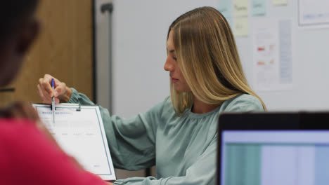 Caucasian-businesswoman-holding-paperwork-talking-to-colleague-in-office-and-smiling