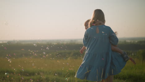 joyful woman in a blue dress holds a little boy in a white shirt, spinning around in a grassy field, with a young boy in yellow behind them.they reach out to touch colorful bubbles floating in the air