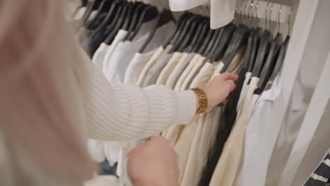 female customer hand in ribbed sweater sleeve with gold chain watch glides along hangers, feeling beige and white shirts, seeking ideal garment inside bright retail rack, demonstrating texture check