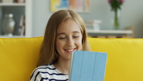 Vista-De-Cerca-De-Una-Adolescente-Rubia-Sentada-En-Un-Sofá-Amarillo-En-Casa-Y-Viendo-Algo-En-Una-Tableta