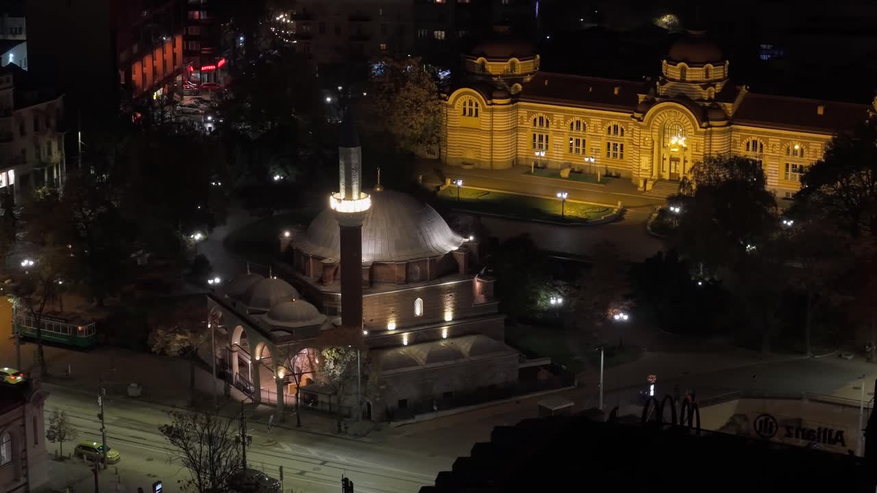 Drone orbit at night around the illuminated Banya Bashi Mosque, revealing the glowing Sofia Regional History Museum in the background
