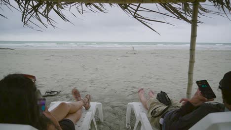 Stormy-ocean-weather-and-couple-relaxing-on-sunbeds-under-gazebo,-dolly-backward-shot
