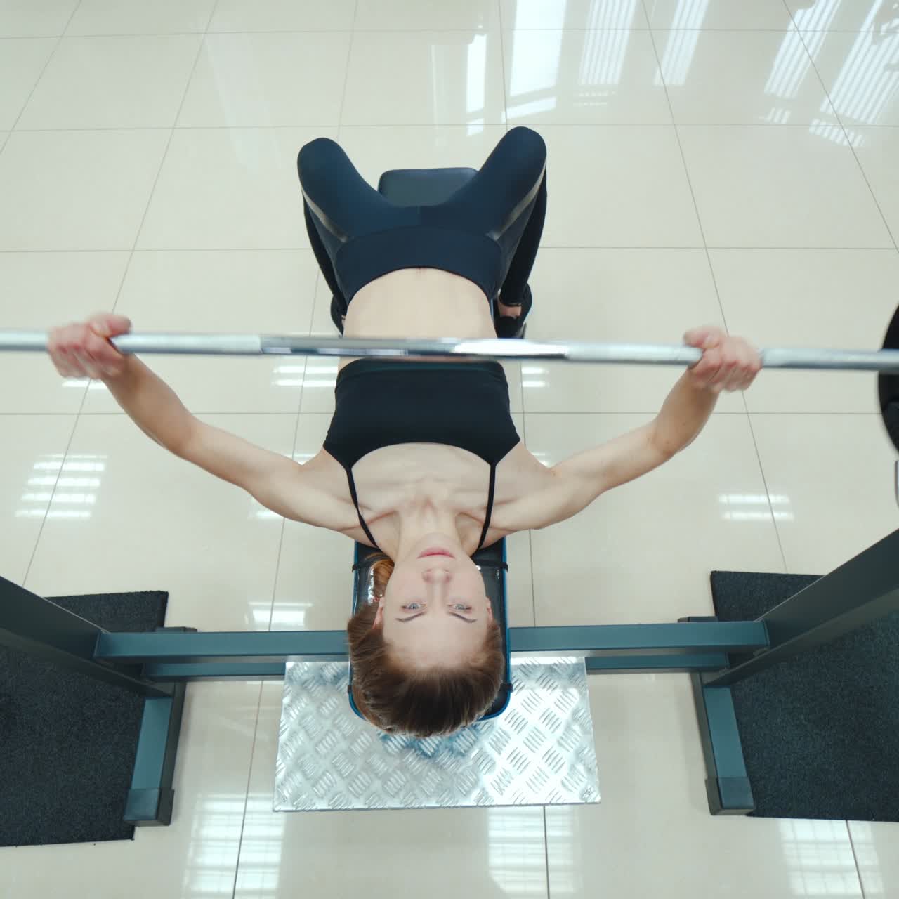 Athletic woman is lying and doing the bench press using the barbell on a white background in the gym. View from above. Endurance exercise