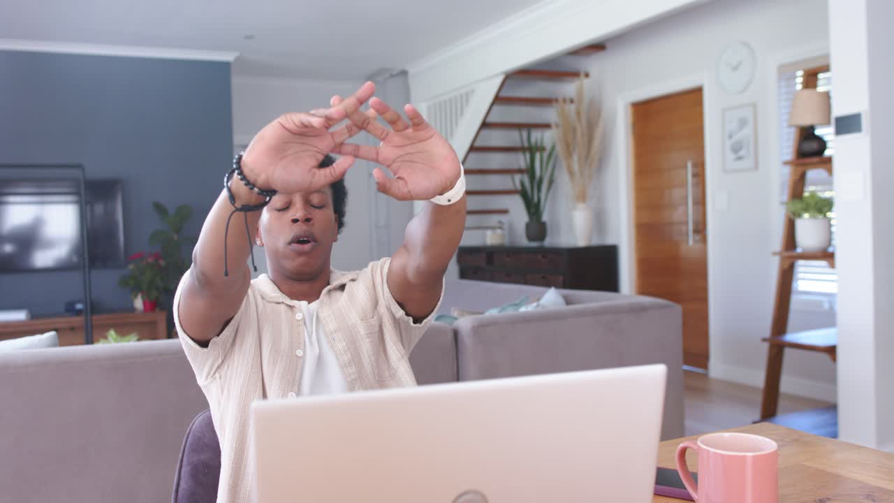 African American man stretching while sitting at home table with laptop and pink mug
