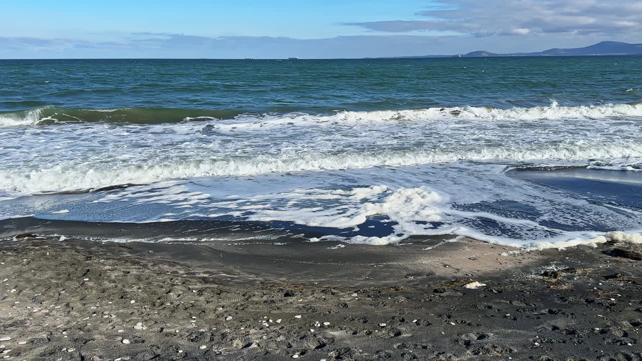 Le onde si infrangono su una spiaggia di sabbia nera vicino all'oceano sotto un cielo azzurro e limpido, con colline distanti visibili all'orizzonte durante il giorno.