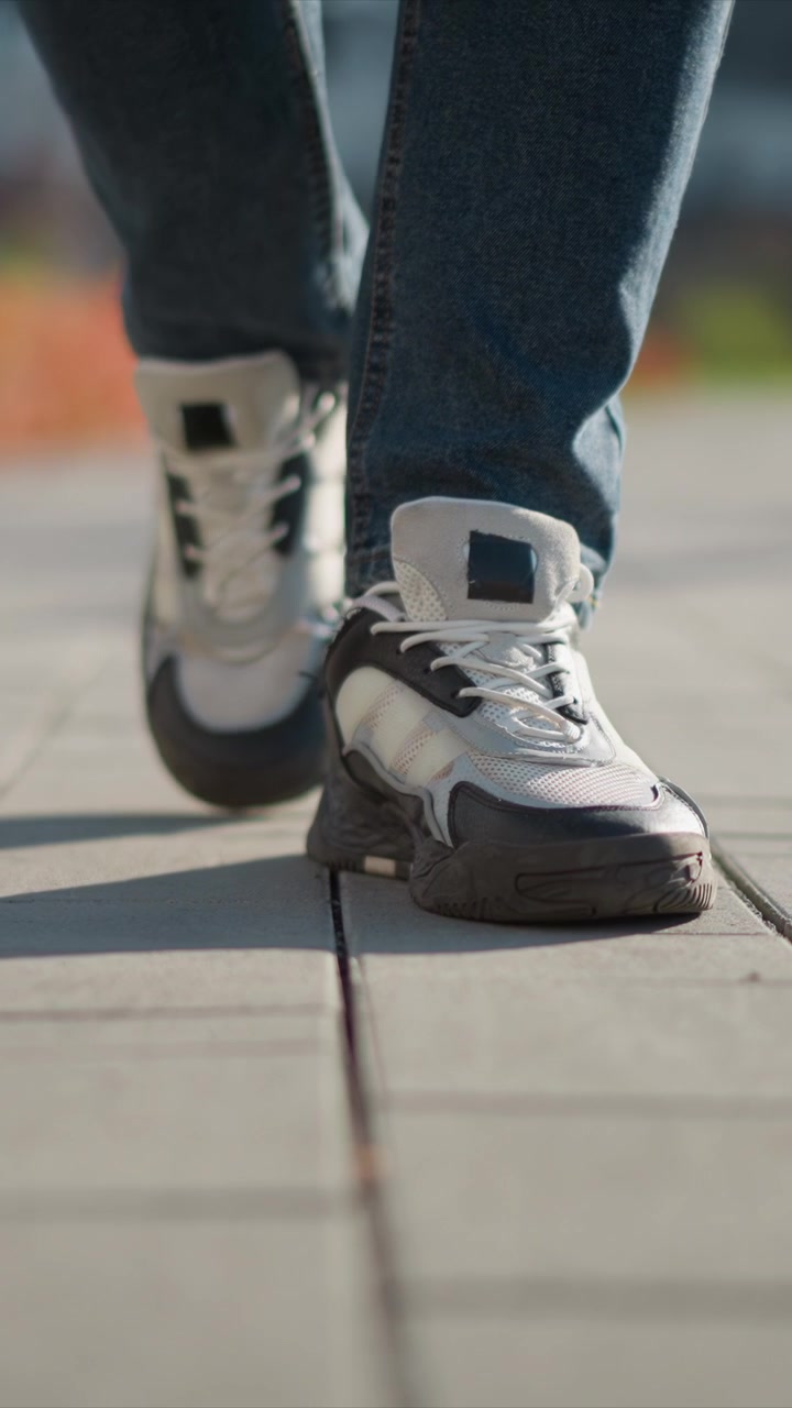 Primo piano dei piedi di una persona che indossa sneakers bianche e nere e jeans blu, mentre cammina su un terreno pavimentato. Le scarpe mostrano una texture dettagliata mentre il soggetto si muove, catturando l'effetto mosso in alcuni fotogrammi.