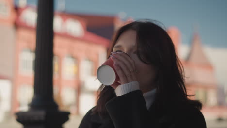 close-up of woman sipping coffee outdoors, fingers adorned with stylish rings, background features blurred red building with clear natural light