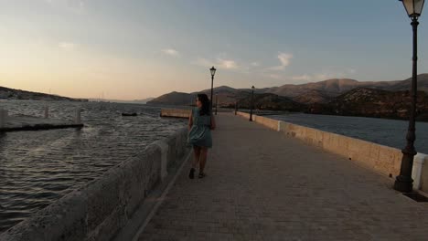 A-girl-in-a-green-dress-walking-down-a-stone-promenade,-looking-at-an-obelisk-and-the-seaside-sunset