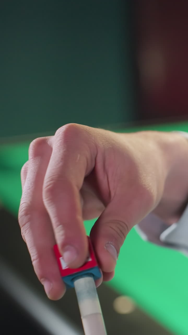 toma de primer plano de la mano con camisa blanca aplicando tiza al palo de señal antes de un juego de billar. mesa verde y fondo borroso añaden profundidad a la escena de preparación enfocada en un entorno interior débilmente iluminado