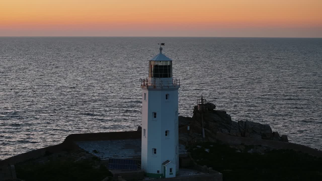 Close drone circle right at Godfrey’s Point Lighthouse silhouette with pink sky and reflective sea, telephoto orbit
