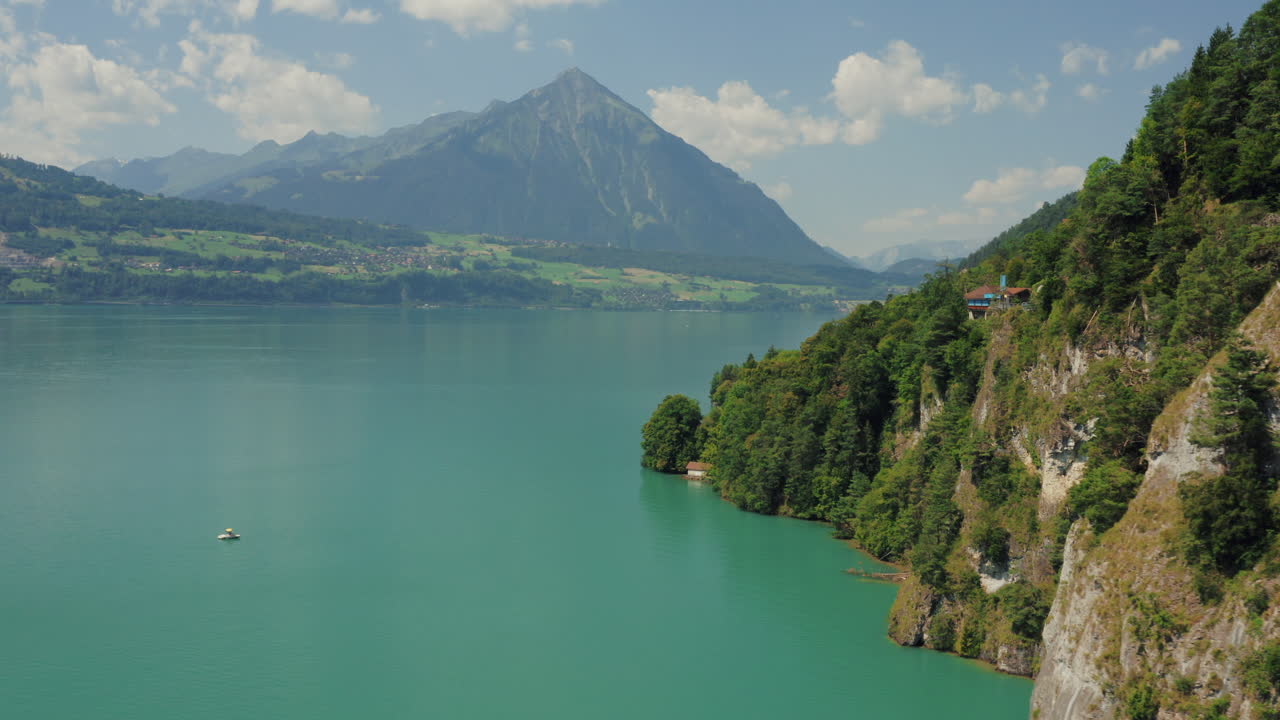 Drone shot of steep cliff revealing turquoise lake with towering mountain in the background. small blue house nestled among the trees. Lone boat drifts on the lake. Switzerland