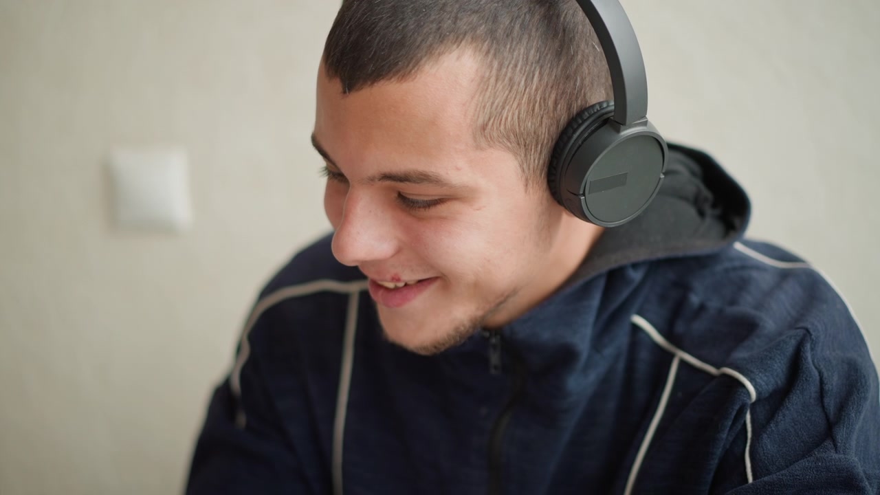 Tampilan dekat pria mengenakan headphone over-ear hitam dan melihat ke bawah. Subjek tetap diam dengan pencahayaan lembut di dalam ruangan, menampilkan nada latar belakang netral dan tanpa gerakan yang terlihat