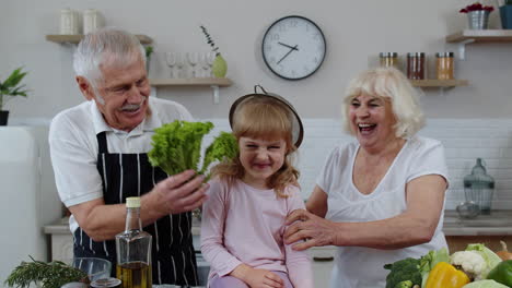 happy vegan senior couple dancing with granddaughter child while cooking vegetables in kitchen