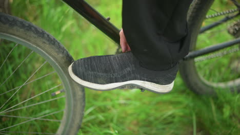 close-up of someone wearing grey sneakers tapping and rotating bicycle pedal in grassy field, placing foot on the pedal, adjust the sneaker before stepping down onto the grassy ground