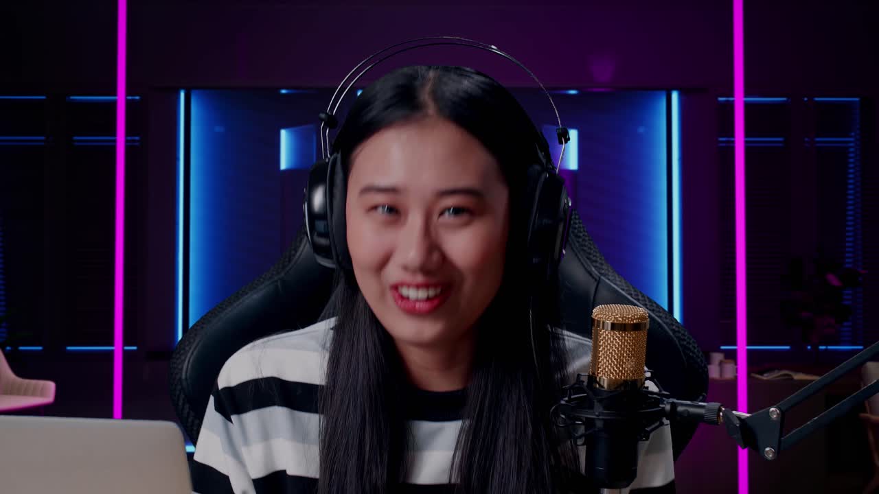 헤드폰과 컴퓨터를 들고 있는 아시아 여성 게이머의 근접 사진. 레트로 형의 네온 조명으로 조명된 방에서 게임을 하는 동안 손을 흔들고 지표손가락을 올린다.