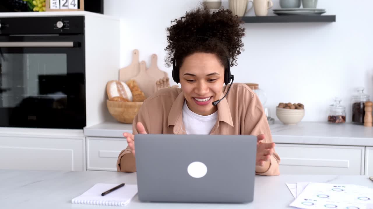 Positive african american girl with headset, freelancer, advisor, student sits at desk in the kitchen, talks via video communication, work or study remotely, gesturing with her hands, smiling happily
