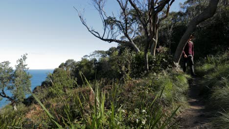 Man-in-hat-walking-on-cliff-top-path,-with-ocean-below-to-the-left-hand-side