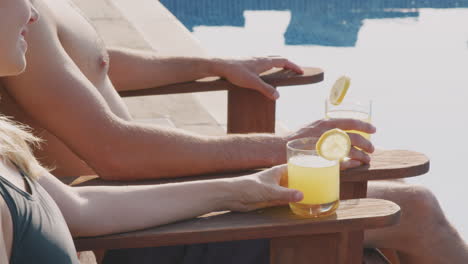 close up of couple with soft drinks on summer vacation sunbathing on loungers by swimming pool