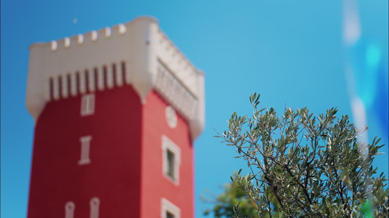 Green tree moving in the wind near the red tower of the Cremat Castle Winery over blue sky