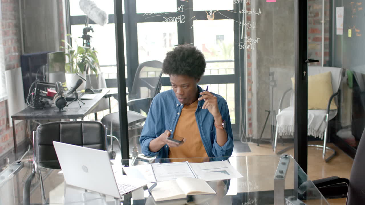Afrikansk-amerikansk afslappet forretningsmand sidder ved skrivebord og bruger smartphone og bærbar computer derhjemme, slowmotion