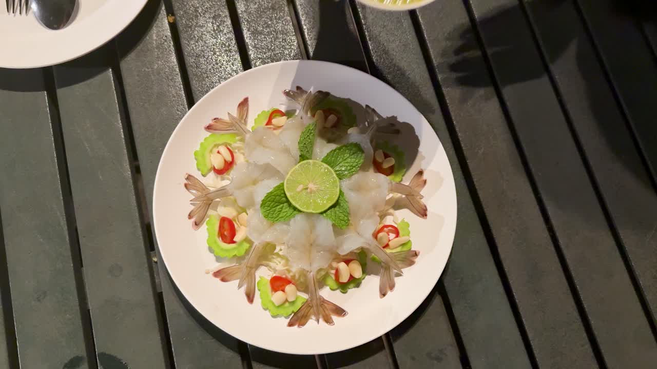 Hand pours green seafood sauce onto raw shrimp platter at outdoor dining table, overhead view