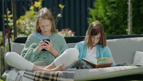 two friends are relaxing on a garden swing. one girl uses a smartphone, another reads a book