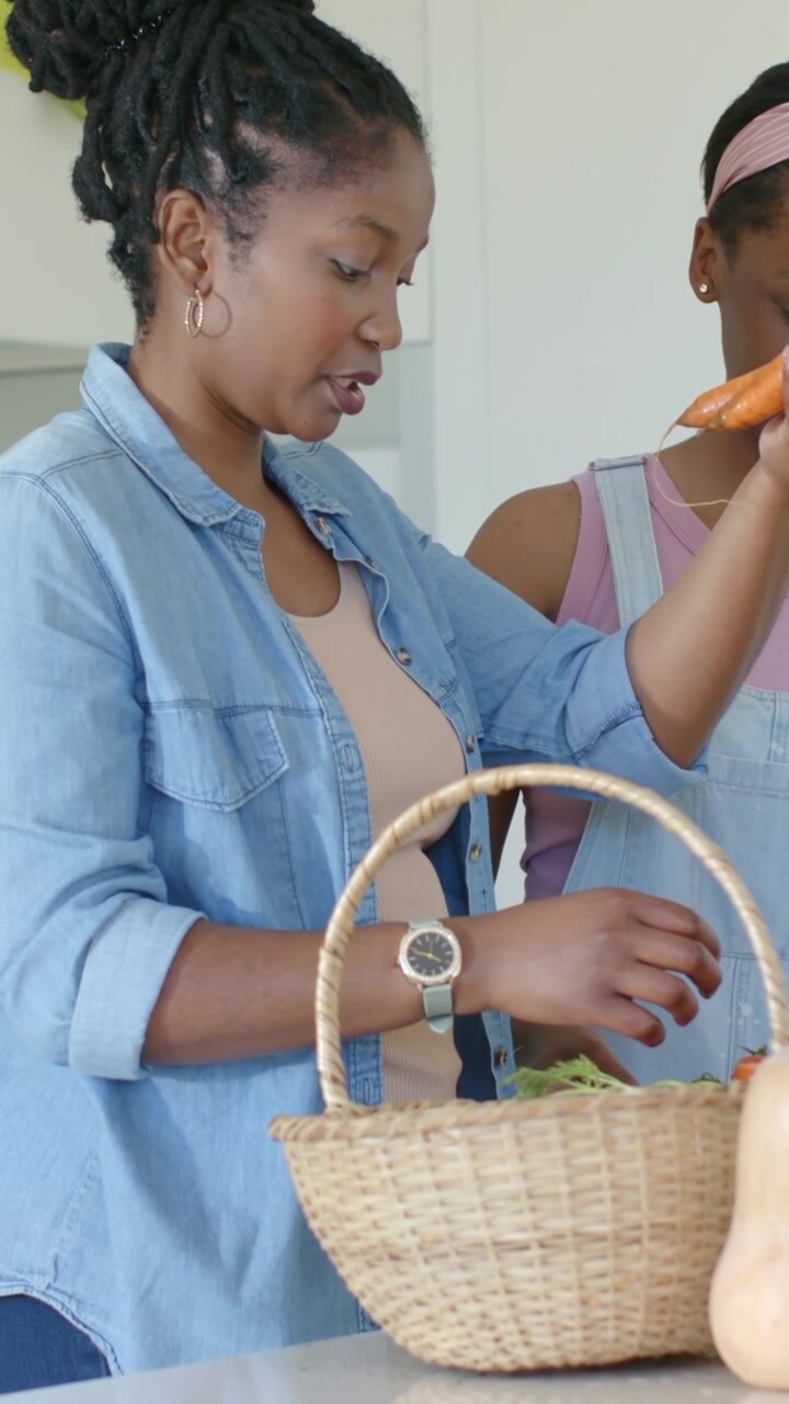 Vertical video: Giving carrot girl overalls urging mom kitchen sorting produce into woven basket