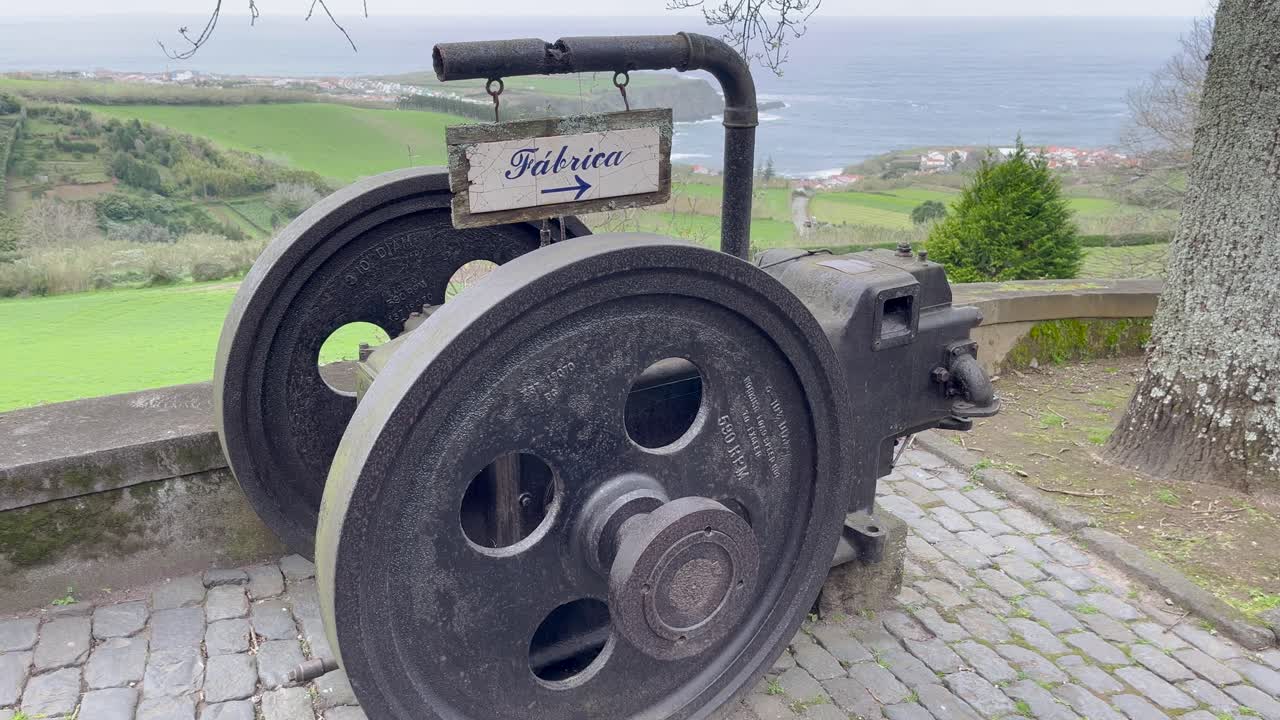 Vintage industrial wheel with a "Fábrica" sign in the Azores, Portugal, set against a scenic countryside and coastal backdrop, highlighting the region's rich industrial heritage and natural beauty.