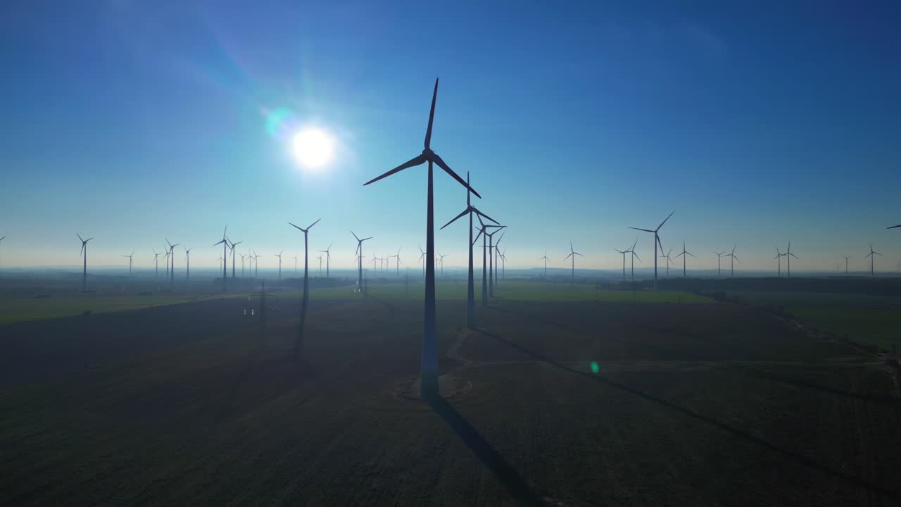 Wind turbines spinning in a vast field, generating sustainable power under a clear blue sky, promoting ecological advancement. Tremendous aerial view ascending raise up drone