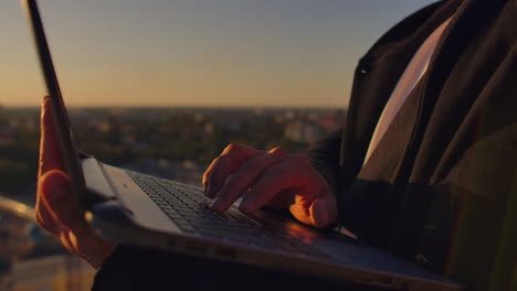 Hacker-using-laptop-on-rooftop-with-city-view-and-forex-chart.-Hacking-and-stats-concept.-A-man-at-sunset-in-slow-motion-writing-software-code-on-a-laptop