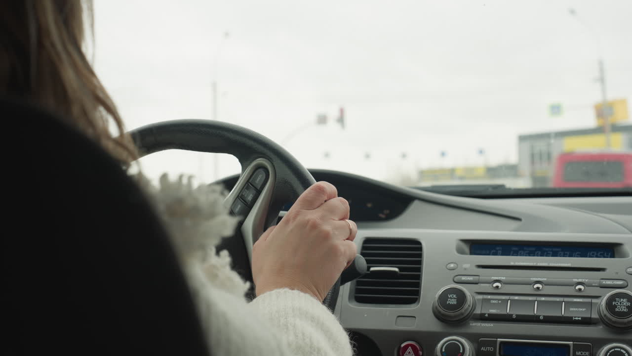 Back view of lady in fluffy white sweater holding steering wheel while patiently waiting at traffic light inside vehicle during daytime with blurred city view and passing cars seen through windshield