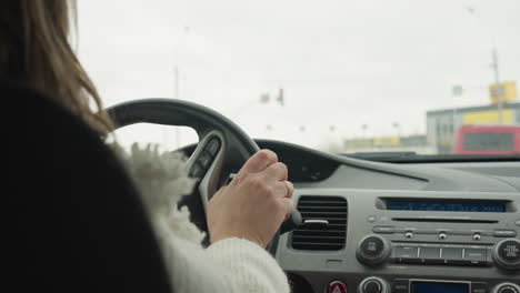 back view of lady in fluffy white sweater holding steering wheel while patiently waiting at traffic light inside vehicle during daytime with blurred city view and passing cars seen through windshield