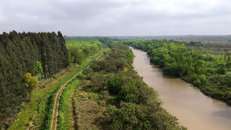 aerial ascend overview of a winding dirt road running alongside a river, cutting through a lush green landscape along river