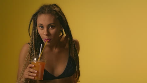 smiling woman drinking orange juice and dancing in studio with yellow wall.