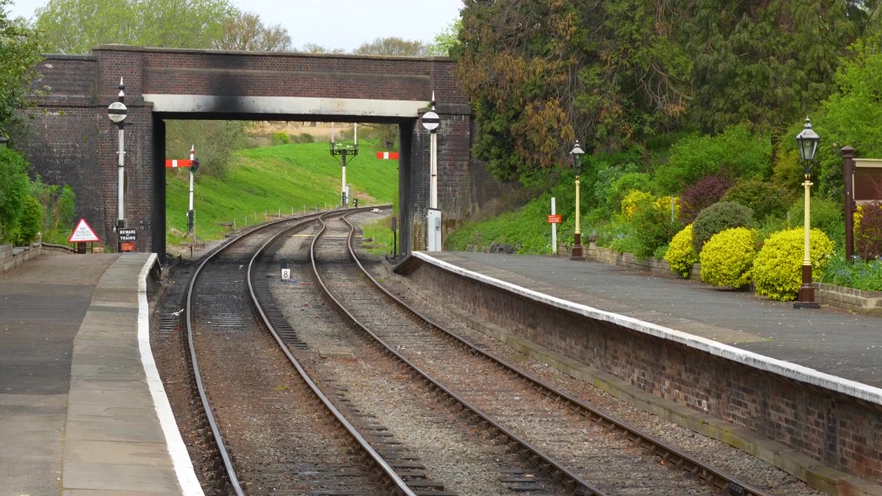 Un'inquadratura ampia di una tranquilla piattaforma di stazione ferroviaria storica con binari paralleli che conducono sotto un ponte di mattoni. Adatto per documentari di viaggio e trasporti.