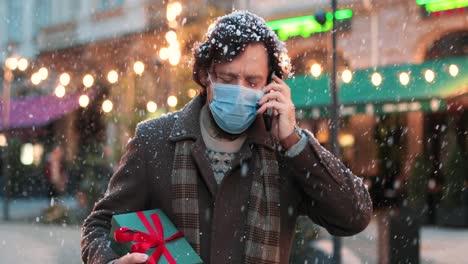 close-up view of caucasian man wearing a face mask talking on phone and holding a present on the street while it‚äôs snowing in christmas