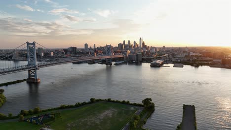 aerial backwards shot of philadelphia skyline, river and famous bridge at sunset. view from camden city shoreline. rising wide shot. marina pier and skyscrapers in distance. film drone shot