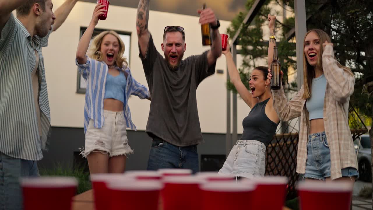 primer plano de un hombre rubio feliz con una botella de cerveza en las manos golpeando una pequeña pelota de tenis de mesa en una taza roja durante una fiesta en el patio de una casa de campo