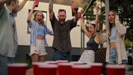 close-up of a happy blond guy with a bottle of beer in his hands hitting a small table tennis ball into a red cup during a party in the courtyard of a country house