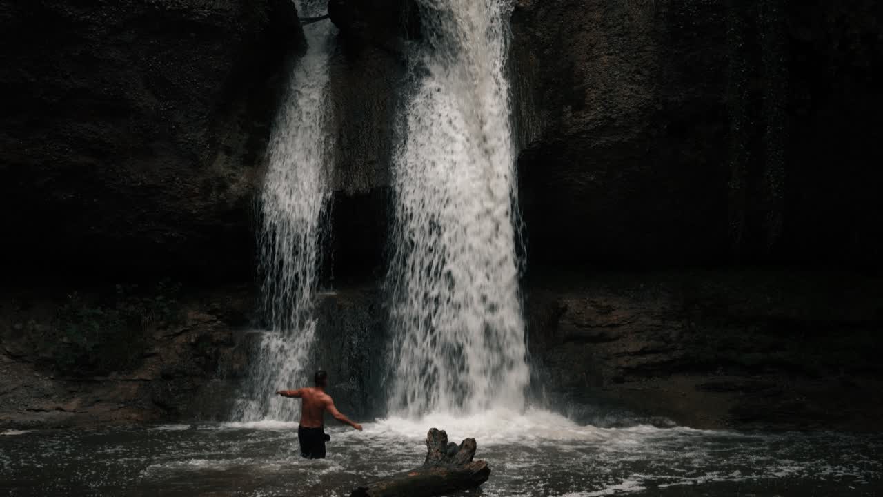 A man runs topless in the water toward a waterfall in a mystical forest. It's cloudy and raining. Epic nature landscape. Shot in slow motion.