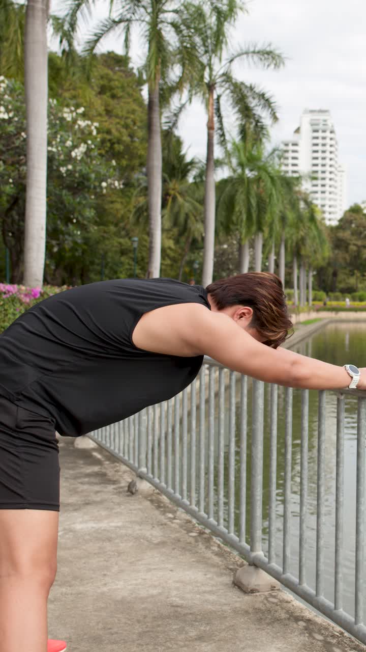 Un hombre asiático en buena forma física hace estiramientos de espalda apoyado en una barandilla de metal en un parque tropical