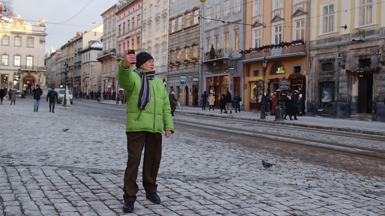 un anciano abuelo turista está haciendo una videollamada en línea con un teléfono inteligente en el centro de la ciudad de invierno